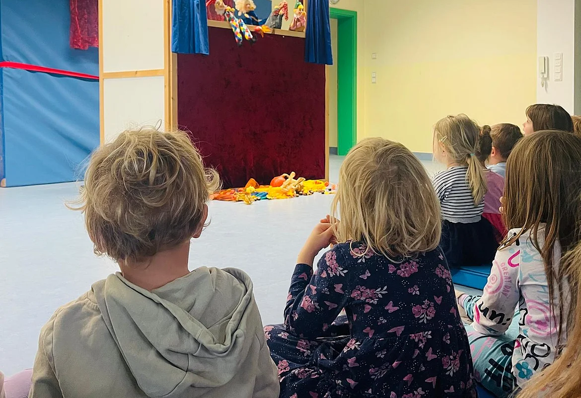 Kinderhaus Thalmassing_Kasperltheater 2025 Mehrere Kinder sitzen im Kindergarten auf dem Boden in einem hellen Raum und schauen gespannt auf ein Kasperltheater. Vor ihnen steht eine kleine Bühne mit rotem Vorhang und blauen Seitenteilen. Hinter der Bühne sind Handpuppen zu sehen, die gerade eine Szene spielen. Auf dem Boden vor der Bühne liegen bunte Stofftiere. Die Stimmung wirkt fröhlich und konzentriert.