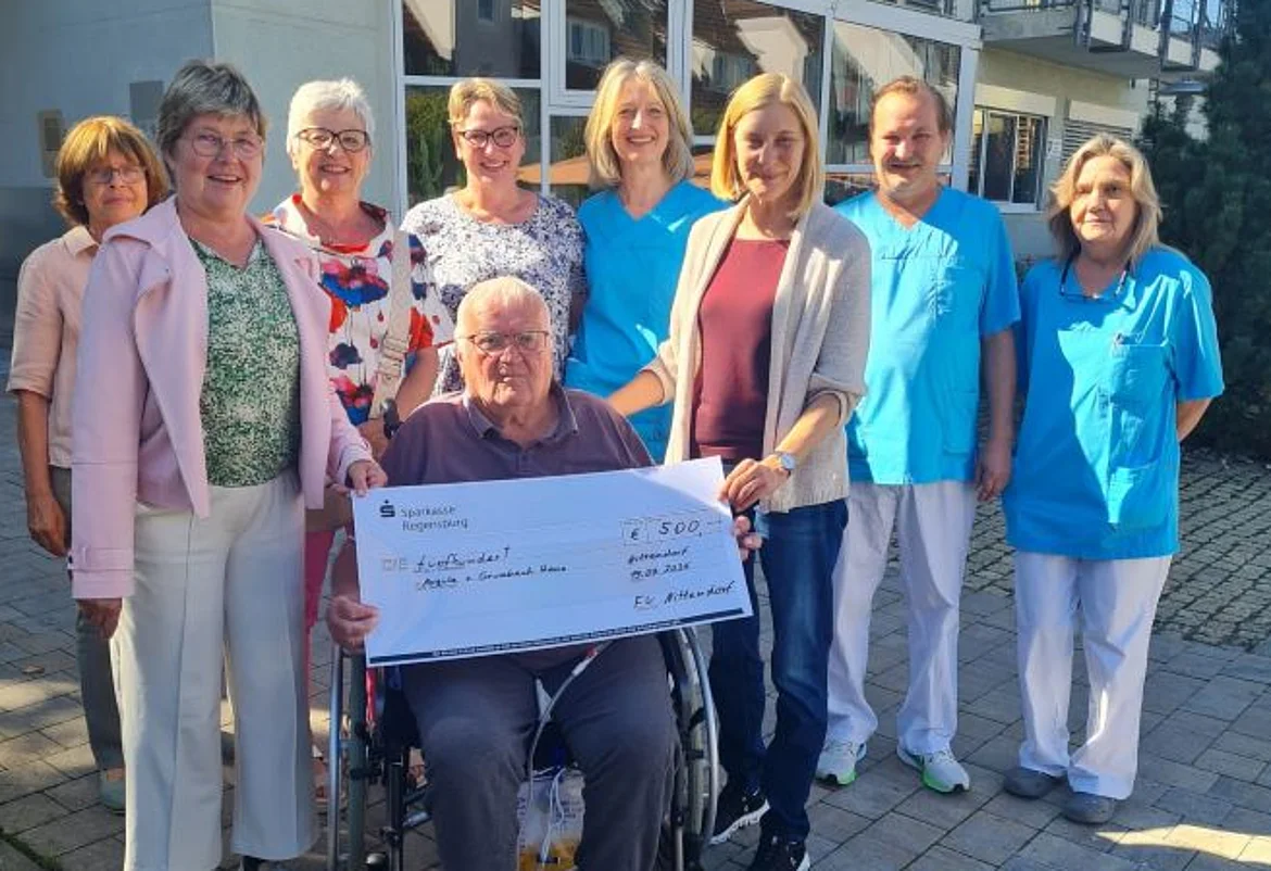 Das Foto zeigt eine Gruppe von insgesamt zehn Personen, die sich vor einem Gebäude versammelt haben. Im Mittelpunkt sitzt ein älterer Mann im Rollstuhl, der einen großen symbolischen Spendenscheck in den Händen hält. Der Scheck ist auf eine Summe von 500 Euro ausgestellt.